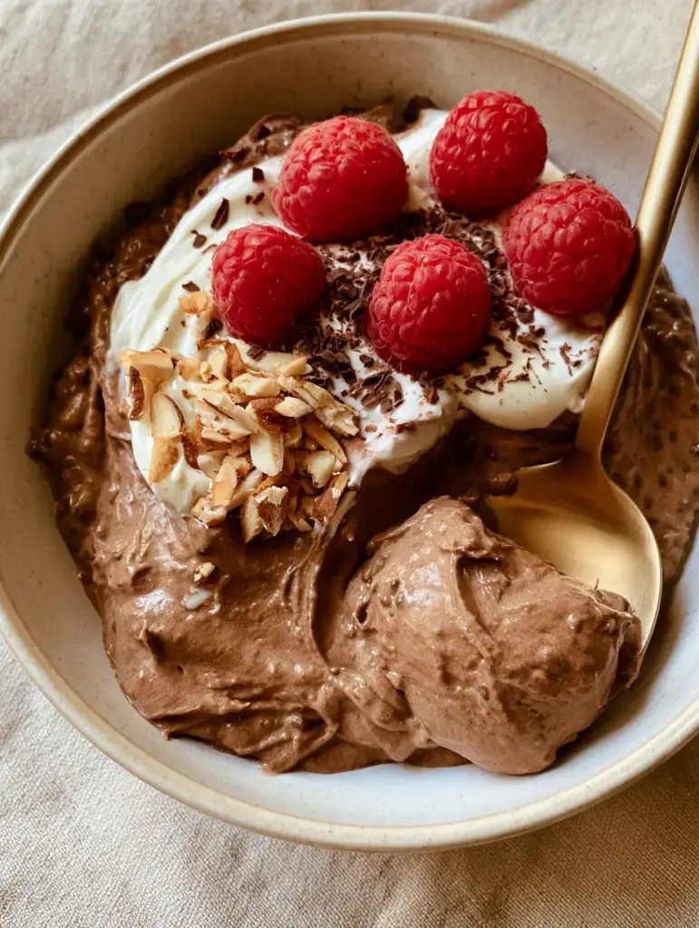 A matte gold spoon lifting a heavy scoop of thick chocolate chia pudding from a beige linen background.