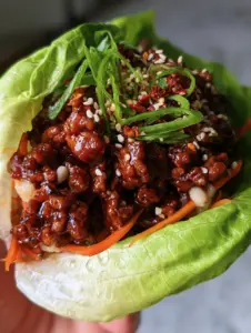 A macro eye-level shot of a single vibrant green butter lettuce leaf cup held by a hand, overflowing with glistening dark brown ground pork crumbles. (pork lettuce wraps)