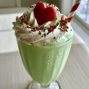 Close-up smartphone photo of a neon green Grinch Vanilla Milkshake with thick whipped cream and a red heart.
